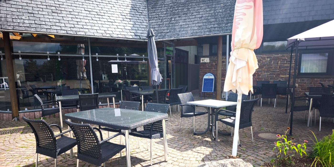 Terrasse vor dem Kiosk am See mit Tischen, Sonnenschirmen und der Tafel mit dem Tagesangebot